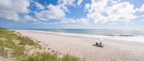 A beach with a wooden deck, grassy dunes, and a clear blue sky.