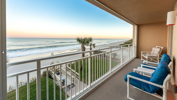 Een balkon met uitzicht op het strand, witte relingen en twee ligstoelen met blauwe kussens.
