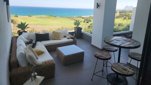 Ein Balkon mit Blick auf den Ozean und einen Golfplatz, ausgestattet mit einer Couch, Hocker und kleinen Tischen.