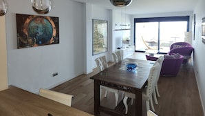 Ein modernes Wohnzimmer mit Essbereich, ausgestattet mit einem Holztisch, weißen Stühlen, einem violetten Sofa und Bildern an den Wänden.