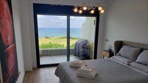 Ein Schlafzimmer mit einem großen Bett, einem Nachttisch, einem Sessel und Blick auf das Meer durch eine Schiebetür.