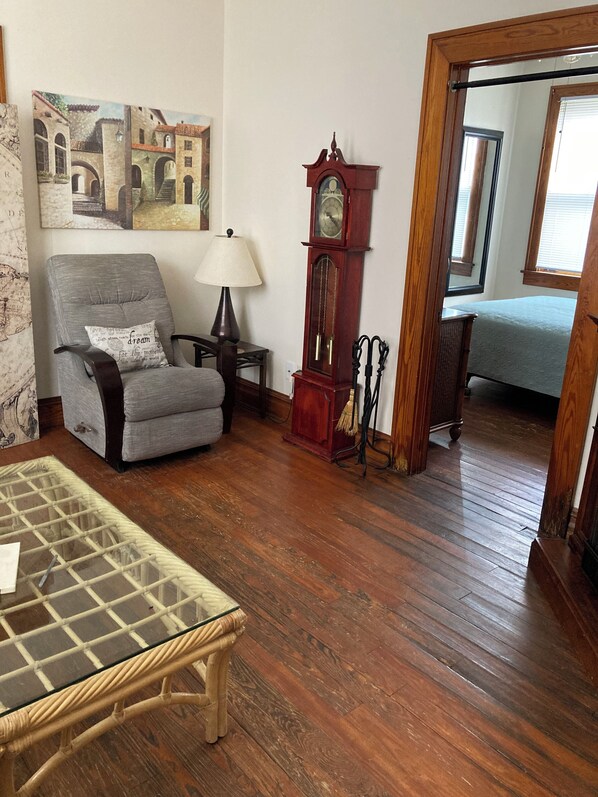 A room with a wooden floor, a grey armchair with a cushion, a glass-top table, a wooden side table with a lamp, a large wooden clock, and a bed in the background.