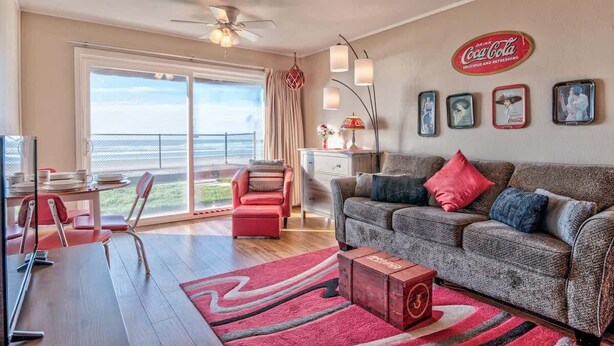 A spacious living room with a large window, a sofa, a dining area, and a Coca-Cola sign on the wall. - Beachfront Suite - Ground Floor - Pool and Sauna! (Lincoln City)