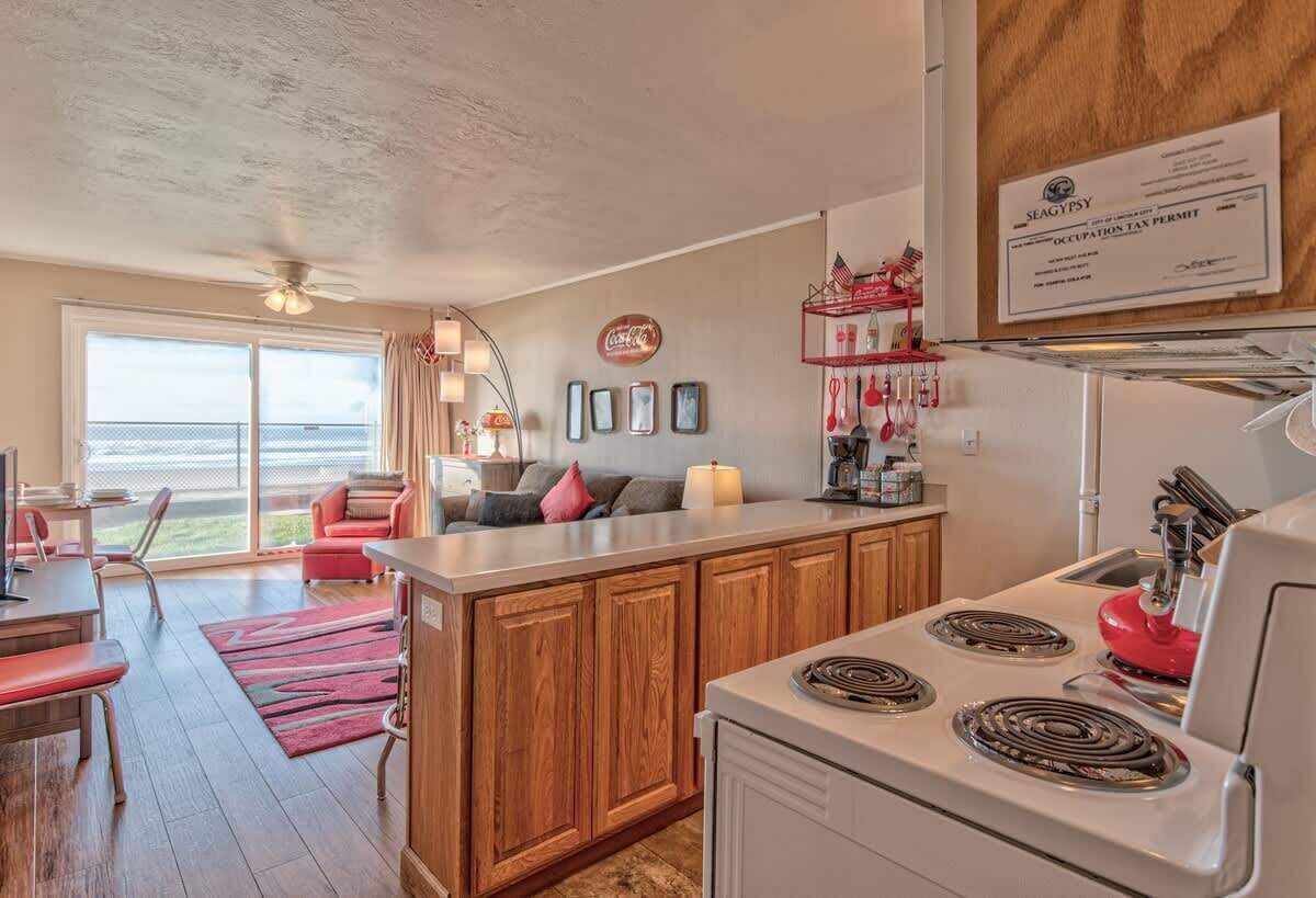 A modern kitchen with wooden cabinets, a central island, and a stove. There is a dining area with a red sofa and a view of the outdoors.