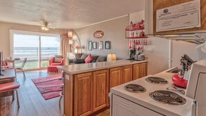 A modern kitchen with wooden cabinets, a central island, and a stove. There is a dining area with a red sofa and a view of the outdoors. - Beachfront Suite - Ground Floor - Pool and Sauna! (Lincoln City)