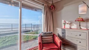 A room with a large sliding glass door leading to a balcony, a red armchair with a patterned cushion, a white dresser with drawers, a Coca-Cola branded lamp, and a decorative vase with flowers. - Beachfront Suite - Ground Floor - Pool and Sauna! (Lincoln City)