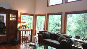 A cozy living room with a wooden cabinet, a glass coffee table, a sofa, and large windows with a view of trees. - "Enchantment"  Medford, Oregon (Medford)