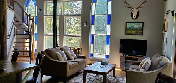 Un salon chaleureux avec un canapé, des fauteuils, une table basse et un téléviseur. De grandes fenêtres aux vitres bleues, un escalier et un décor mural représentant un crâne de cerf complètent l’ensemble.
