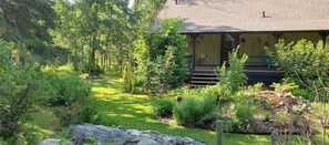 Une maison dotée d’un porche abrité, entourée d’un jardin luxuriant où poussent différentes espèces de plantes et où se trouve un petit étang.