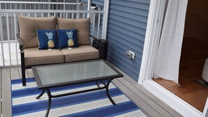 Een overdekt terras met een beige bank, twee blauwe kussens met ananasdessins, een tafel met glazen blad en een blauw-wit gestreept vloerkleed.