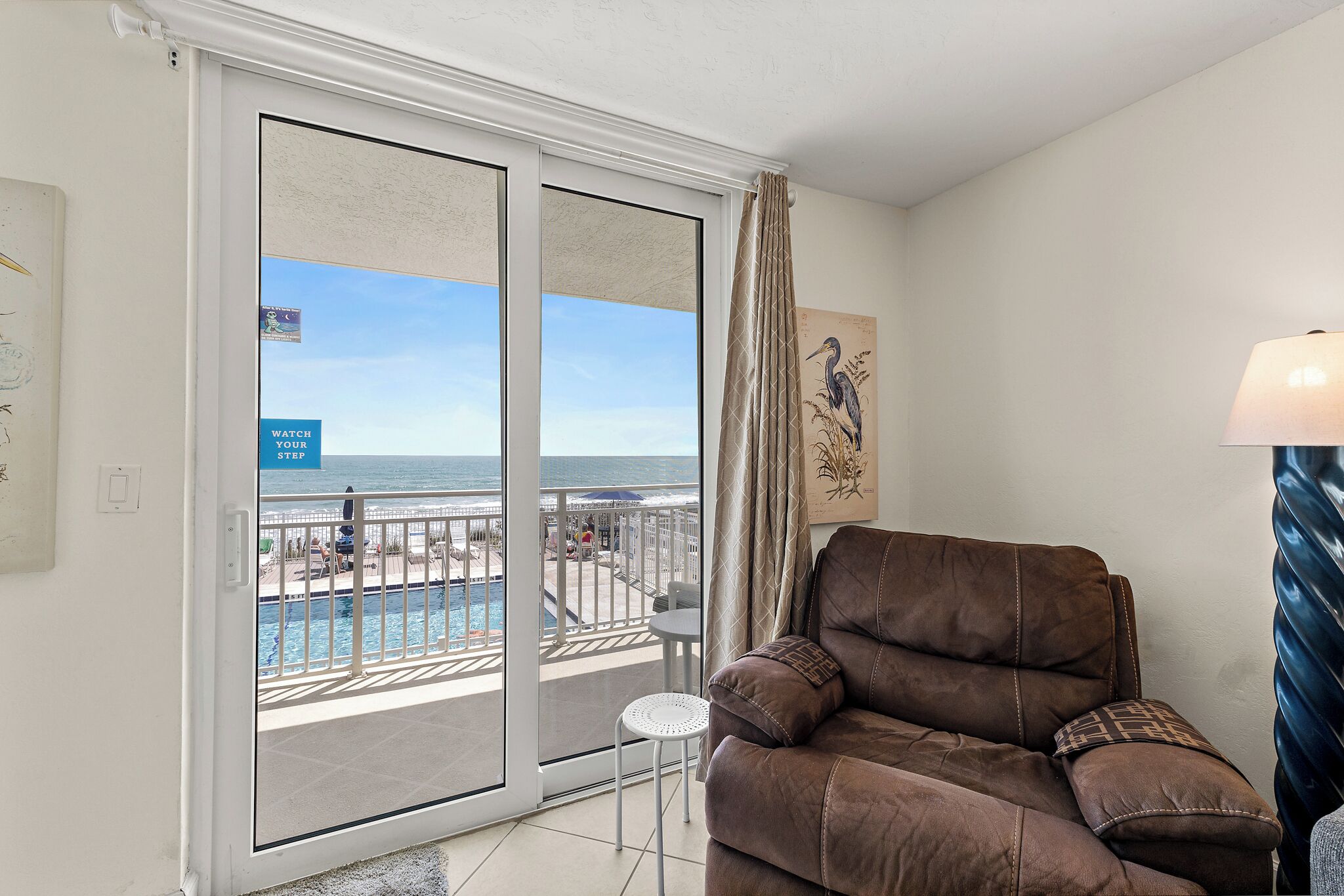 A room with a brown leather sofa, a small white table, a lamp, a painting, and a sliding glass door leading to a balcony with a pool and a view of the ocean.
