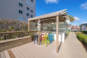 Une terrasse en bois avec des tabourets colorés, une pergola et un panneau indiquant : « À manger. »