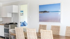 A dining area with wicker chairs, a wooden table, and a kitchen with white cabinets and a stove. - Mollymook At The Beach (Mollymook Beach)