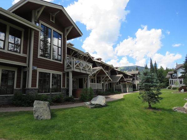 A large, multi-story building with a prominent balcony area, stone foundations, and a well-maintained lawn with a few trees and rocks.