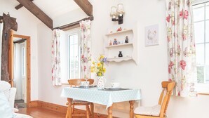Un coin repas avec une table et des chaises en bois, des rideaux à motifs floraux et une étagère ornée d’objets décoratifs.