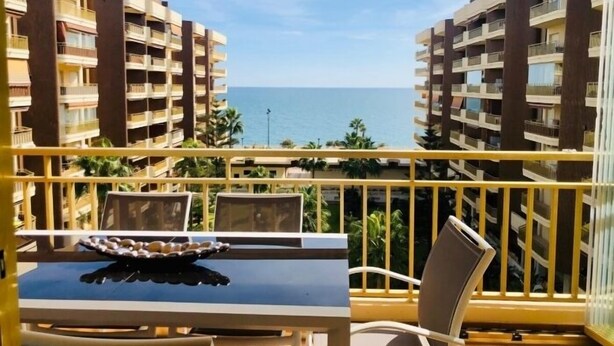 A balcony with a table and chairs, overlooking a sea view with buildings in the background.