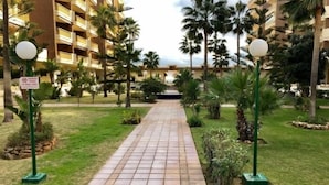 Un chemin pavé mène à un bâtiment doté de balcons, qui est entouré de palmiers et de verdure.