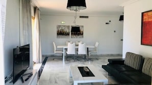 Un salon moderne doté d’un téléviseur à écran plat, d’un canapé noir, d’une table basse blanche et d’un coin repas avec une table et des chaises blanches.