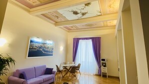 Ein Wohnzimmer mit violettem Sofa, Holzboden, einem Esstisch mit Stühlen und einer Decke mit dekorativen Mustern.