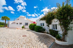 A white building with a tiled courtyard, a palm tree, and a small bush with pink flowers.