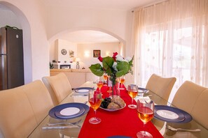 A dining area with a red tablecloth, white plates, and glasses, a vase with flowers, and a kitchen in the background.