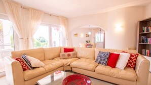 A living room with a beige sectional sofa, a glass coffee table, and a bookshelf. - Casa Alegria · This villa is perfect for a family (Lagoa)