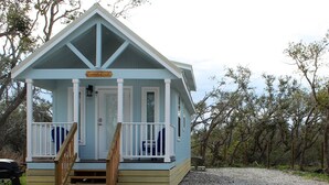 A small, light blue cabin with a porch and wooden steps, surrounded by gravel and trees. - Coast Oak -Beautiful New Tiny Cottage - Perfect Family Getaway (Aransas Pass)