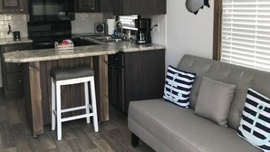 A modern kitchen with dark wood cabinets, a built-in island with bar stools, a gray sofa with striped pillows, and decorative fish wall art. - Coast Oak -Beautiful New Tiny Cottage - Perfect Family Getaway (Aransas Pass)