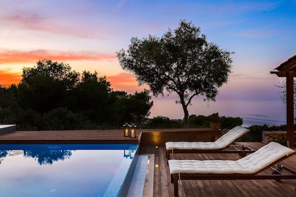 A poolside area with lounge chairs and a wooden deck, overlooking a sunset.