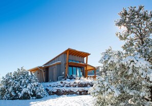 Eine moderne Holzhütte mit großer Veranda und Steintreppe, umgeben von schneebedeckten Bäumen.