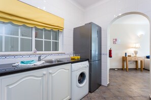 A compact kitchen with a built-in washing machine, a refrigerator, and a window with a yellow curtain.