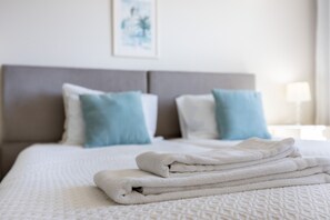 Un lit bien fait, avec du linge de lit blanc et des serviettes pliées, une tête de lit grise et deux oreillers, un bleu et un blanc, dans une chambre d’hôtel.