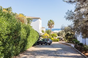 A cobblestone street with a parked SUV, surrounded by lush greenery and a white building with a tiled roof.