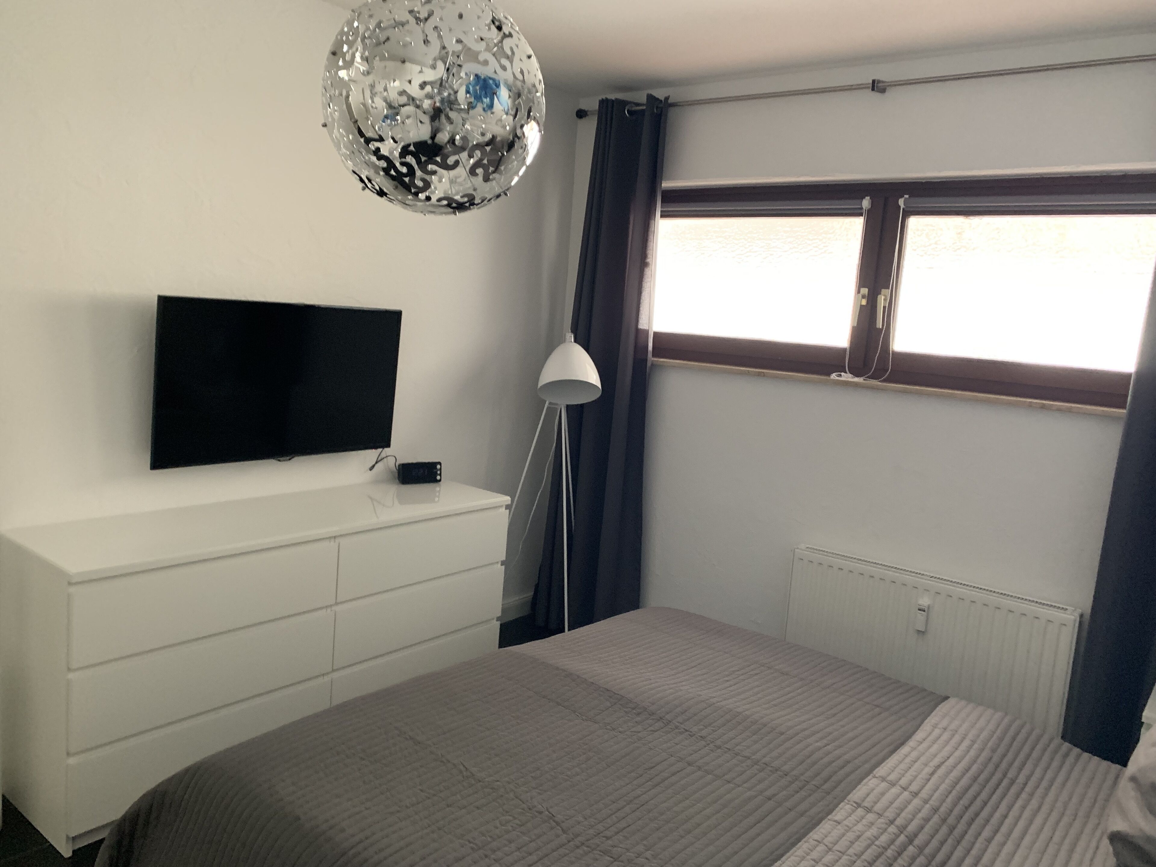 A bedroom with a bed, a TV on a white dresser, a lamp, and a window with curtains.