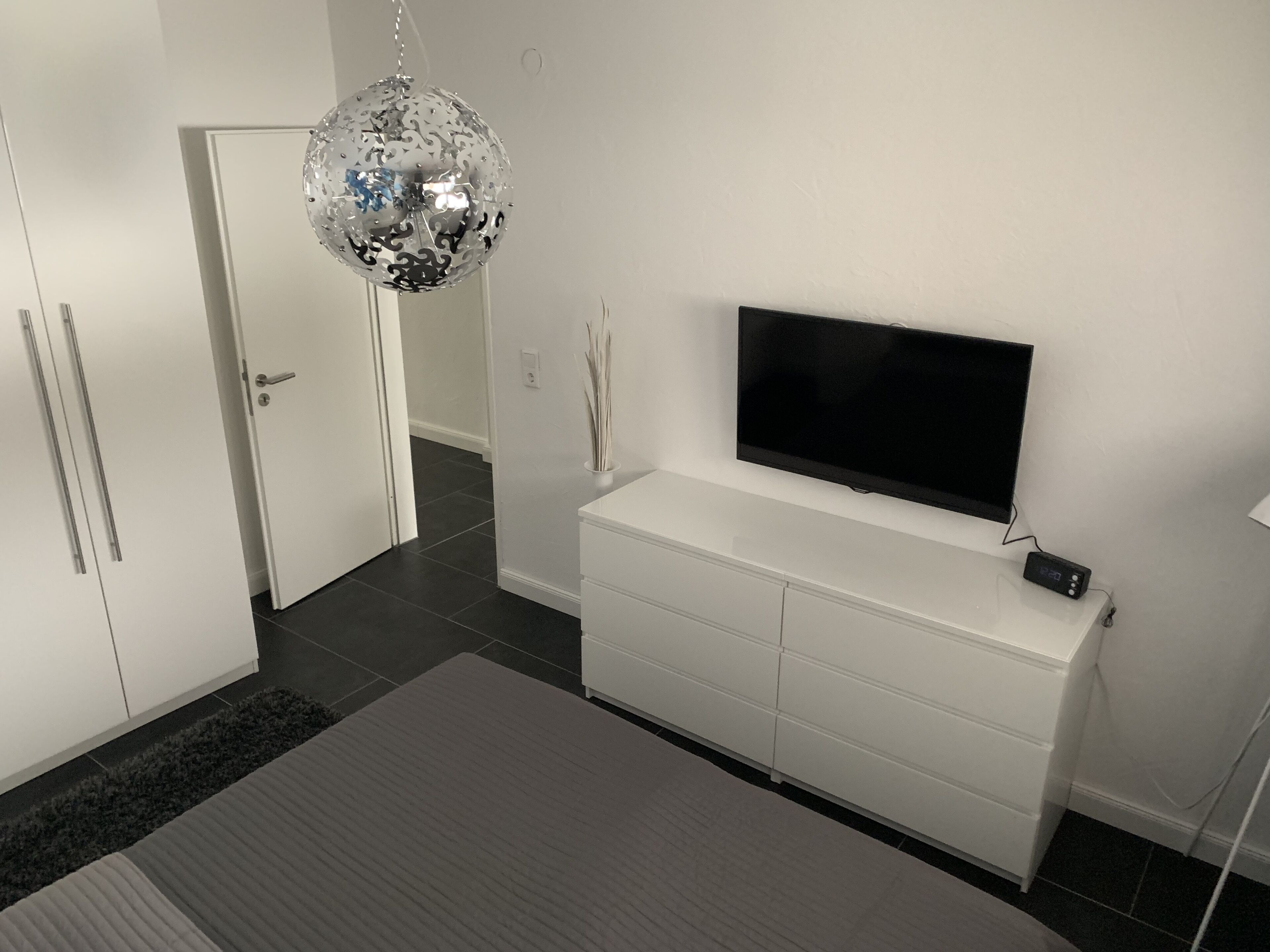 Un dormitorio moderno con televisor, armario blanco y una lámpara decorativa.