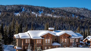Un grand chalet doté de nombreuses constructions en bois, entouré d’arbres couverts de neige et adossé à un paysage montagneux.
