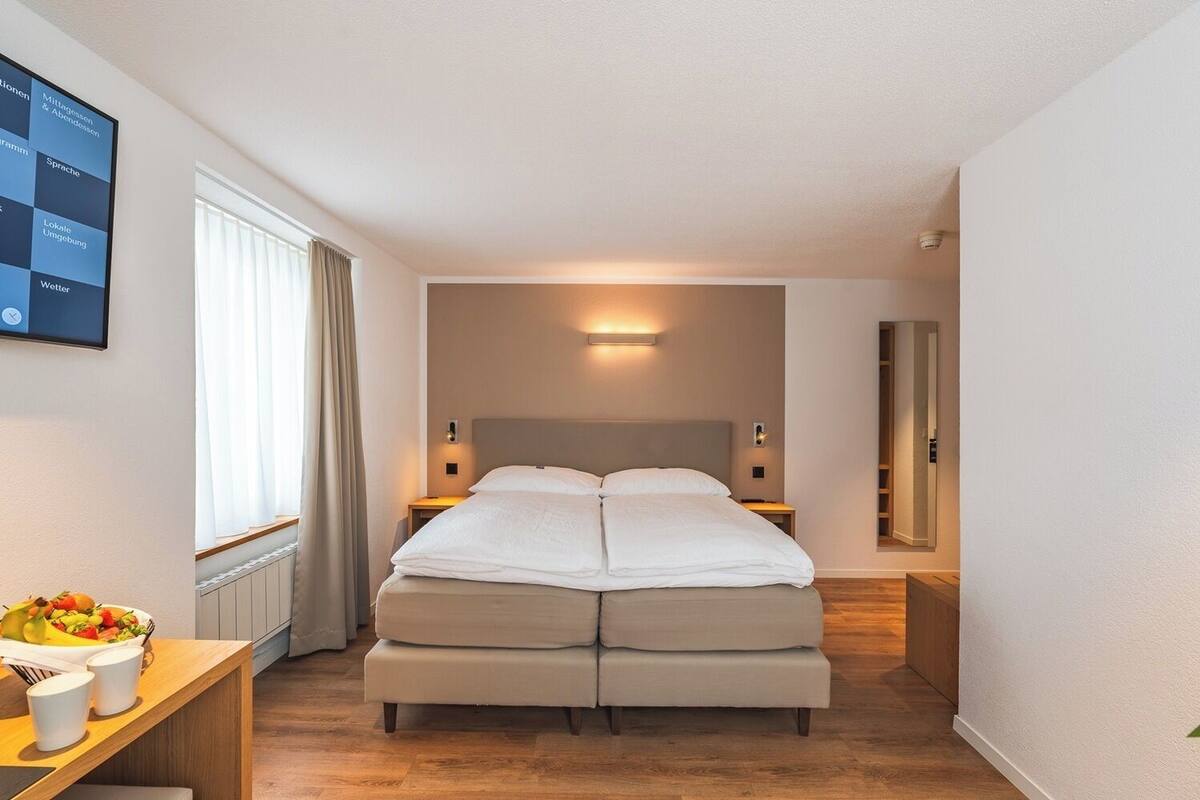 A modern hotel room with a large bed, bedside tables, a small desk with a fruit bowl, and a wall-mounted TV displaying a menu.
