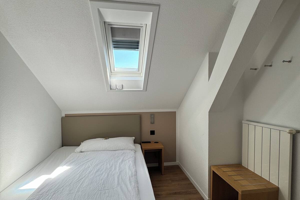 A bedroom with a sloped ceiling, a skylight, a bed, a bedside table, and a bench.
