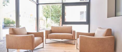 Un interior moderno con tres sillones de cuero dispuestos alrededor de un piso de madera, amplias ventanas que ofrecen vistas al exterior.