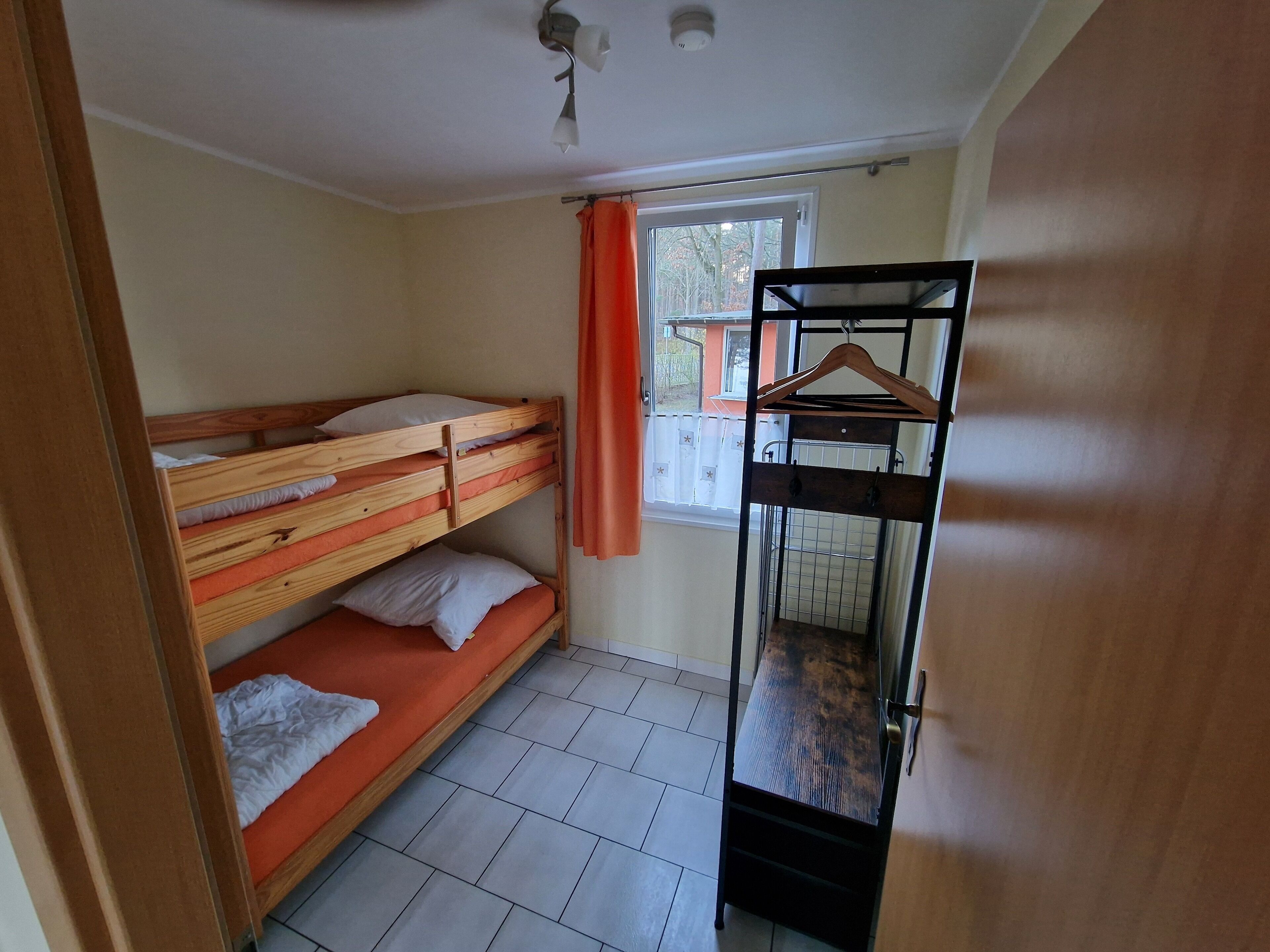 A dormitory room with bunk beds, a window with curtains, a metal rack, and a tiled floor.