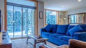 Una sala de estar con un sofá azul, una mesa de centro de madera y un ventanal con vistas a árboles nevados.