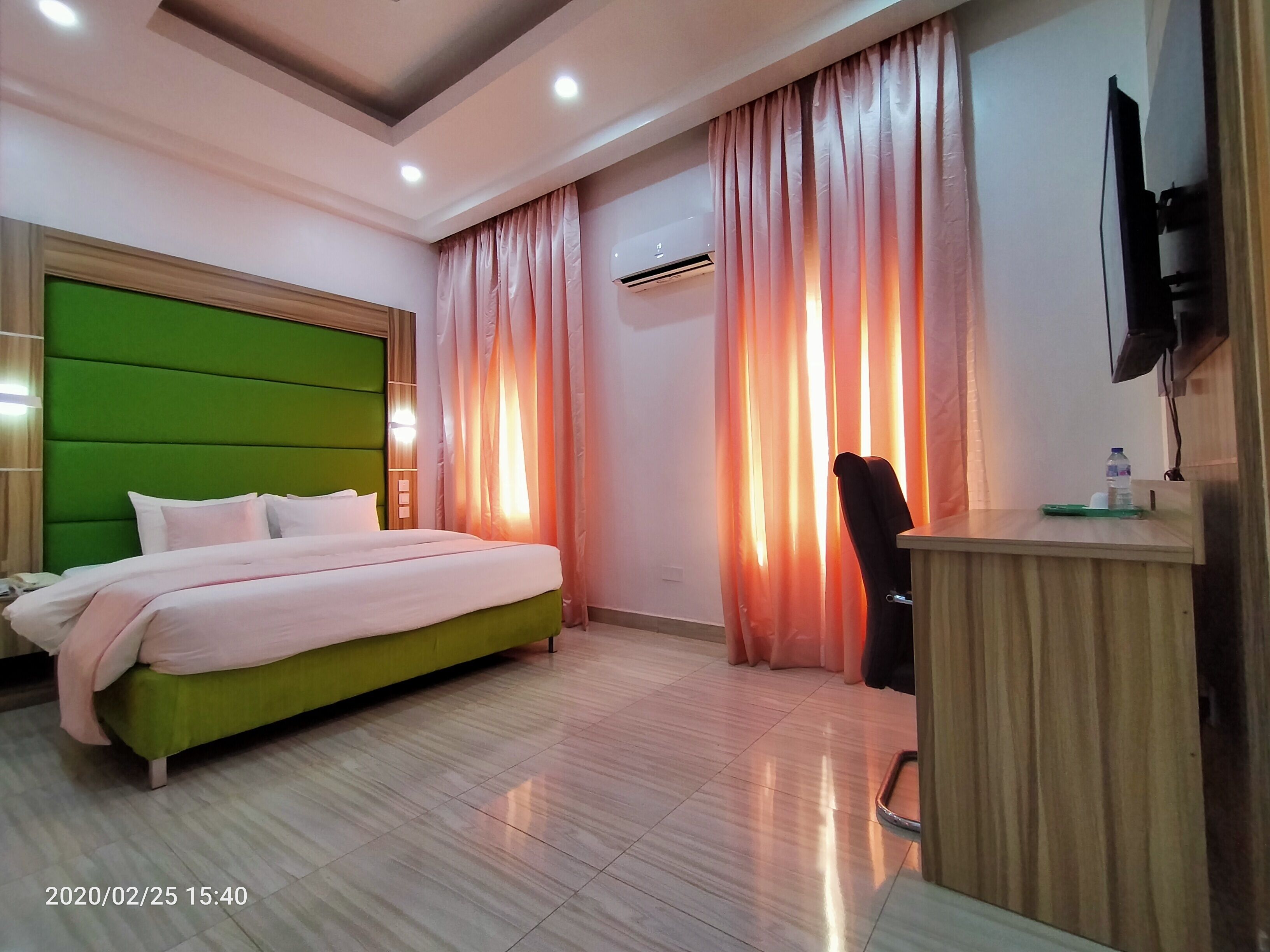 A hotel room with a green headboard, a bed with white bedding, a desk with a chair, and an air conditioning unit.