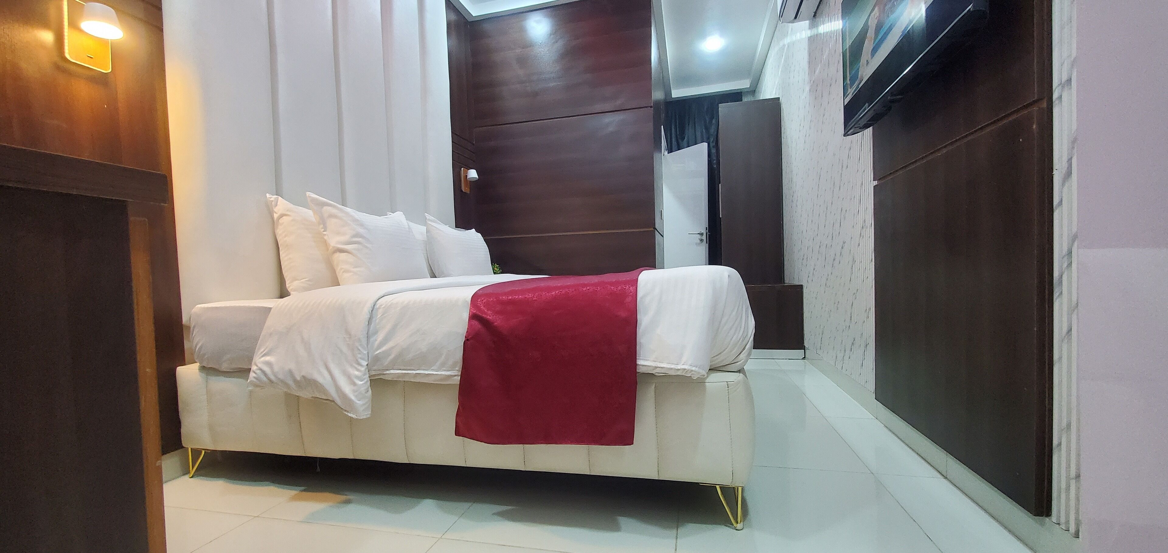 Quarto com cama, roupa de cama branca, manta vermelha, guarda-roupa de madeira e televisão fixada na parede.