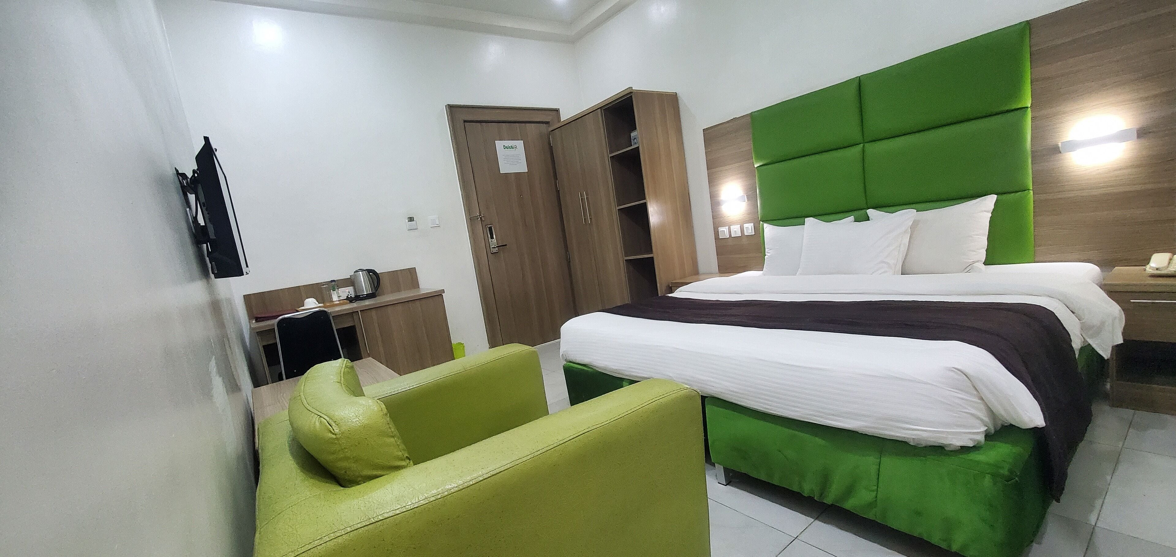 Une chambre d’hôtel comprenant un lit, un fauteuil recouvert d’un tissu vert, une armoire en bois et un bureau avec une bouilloire.