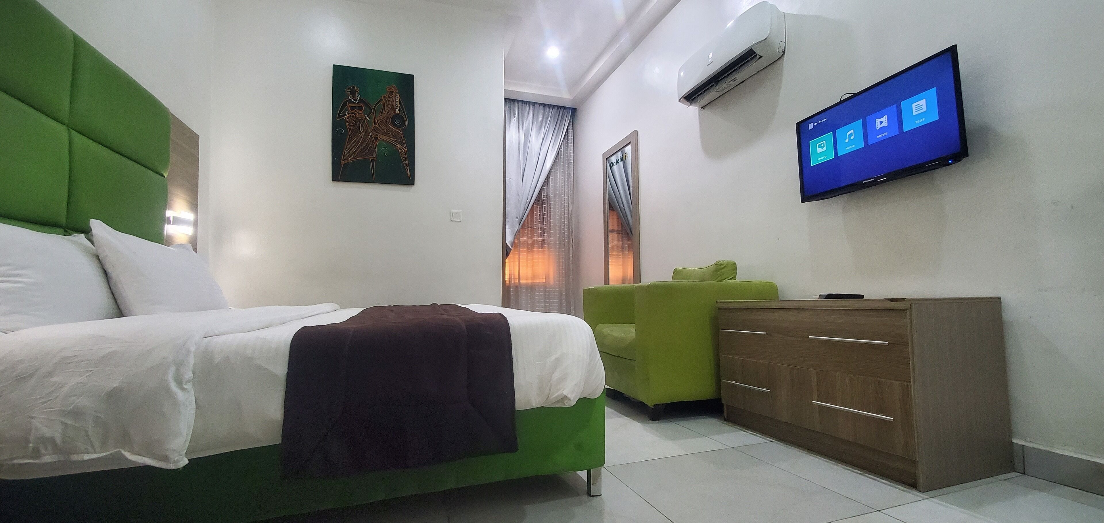 Habitación de hotel con una cama, un sillón verde, un aparador de madera y un televisor de pared mostrando una interfaz de Windows.