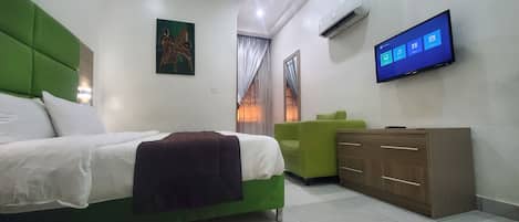 Habitación de hotel con una cama, un sillón verde, un aparador de madera y un televisor de pared mostrando una interfaz de Windows.