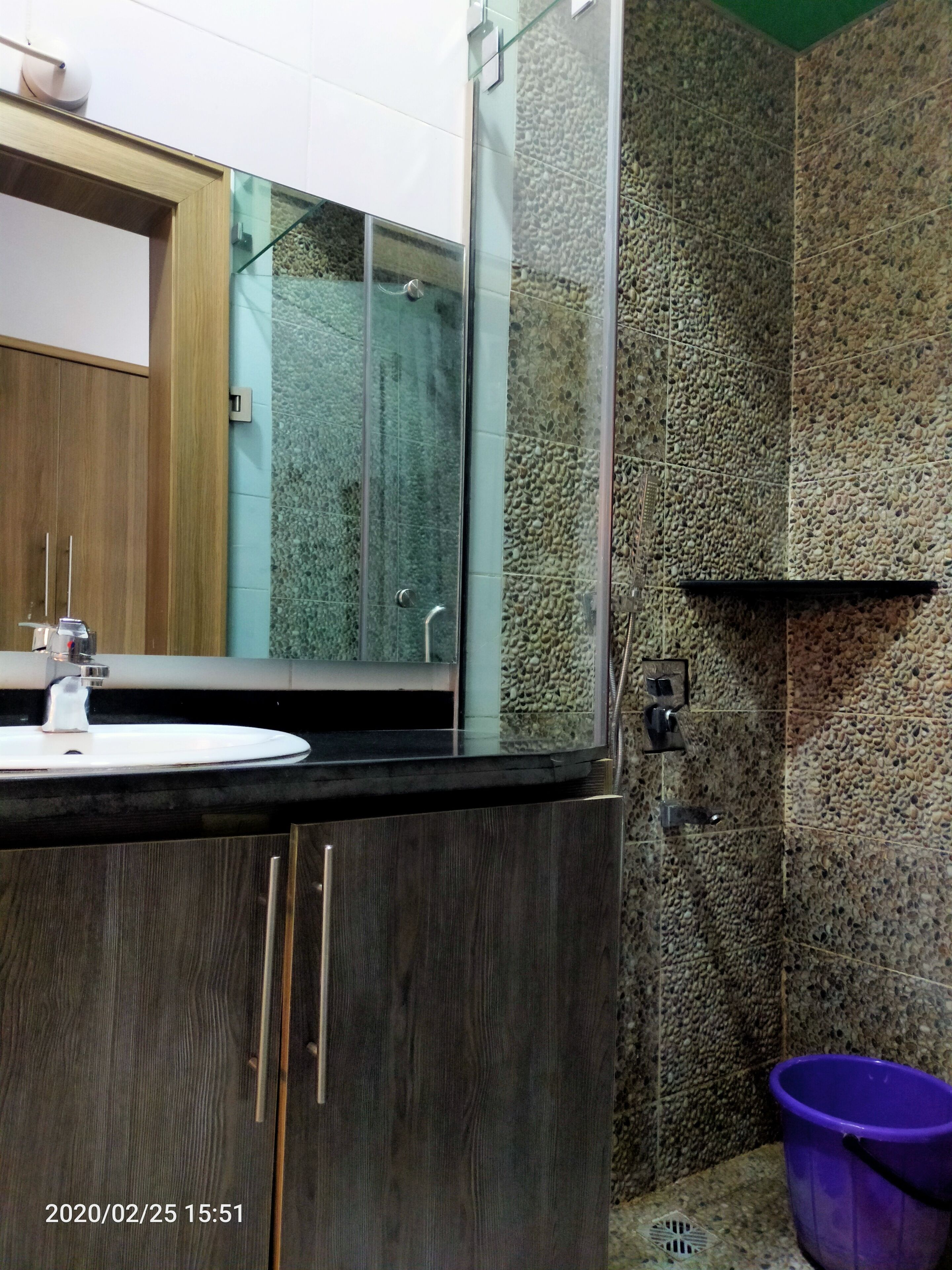 Ein Badezimmer mit einem hölzernen Waschtisch, einem weißen Waschbecken, einer Duschkabine aus Glas und einer Wand mit Kieselstein-Struktur.