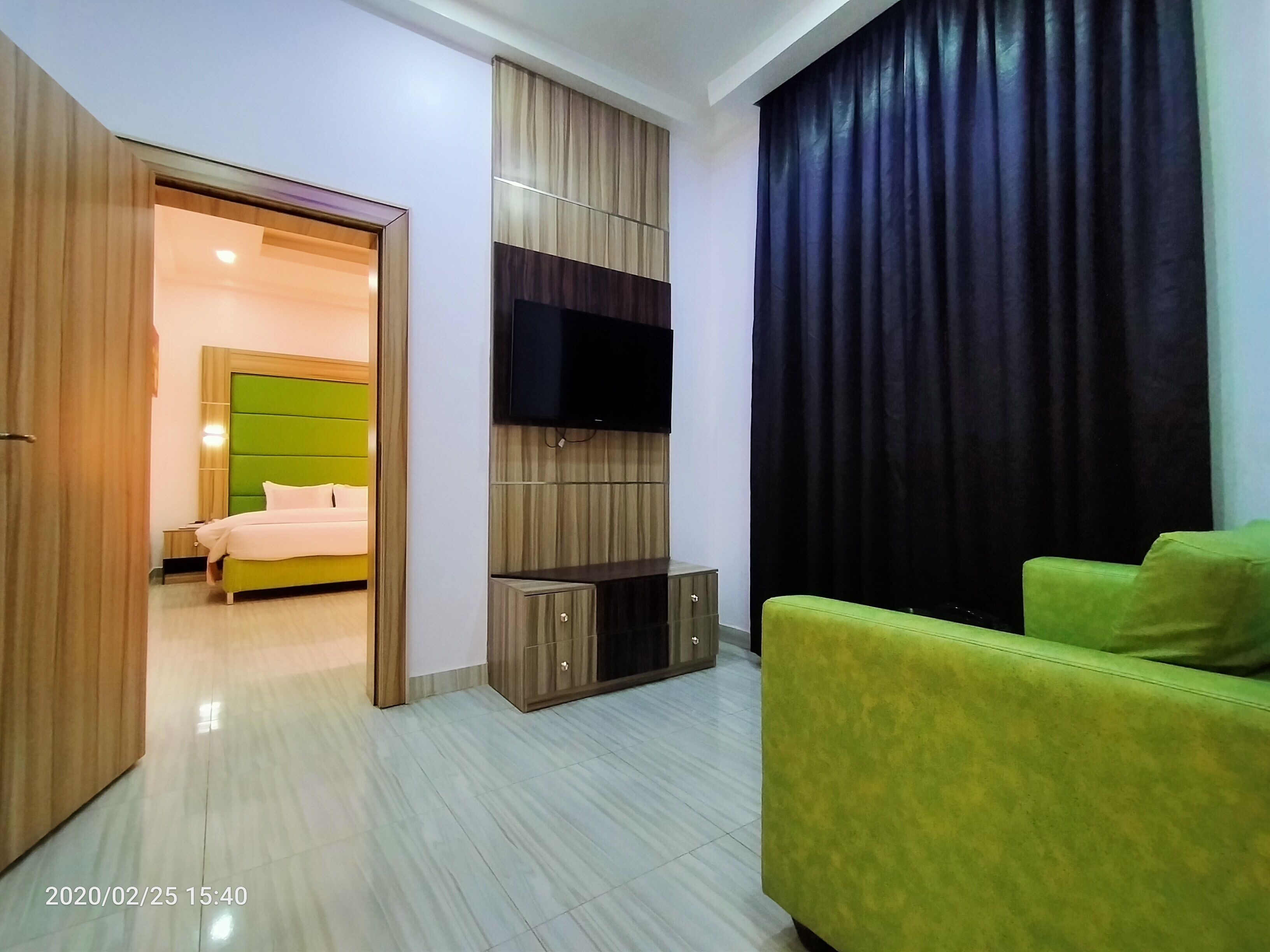 A modern hotel room with a green sofa, a flat-screen TV mounted on a wooden panel, a bed with a green headboard, and a large window with dark curtains.