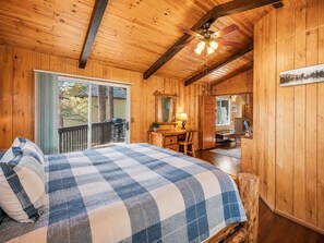 A wooden cabin interior with a bed, a dining area, and a view through a sliding door. - Moonridge Manor - Close to Slopes, Hot Tub, Game Room, Pool Table (Big Bear Lake)