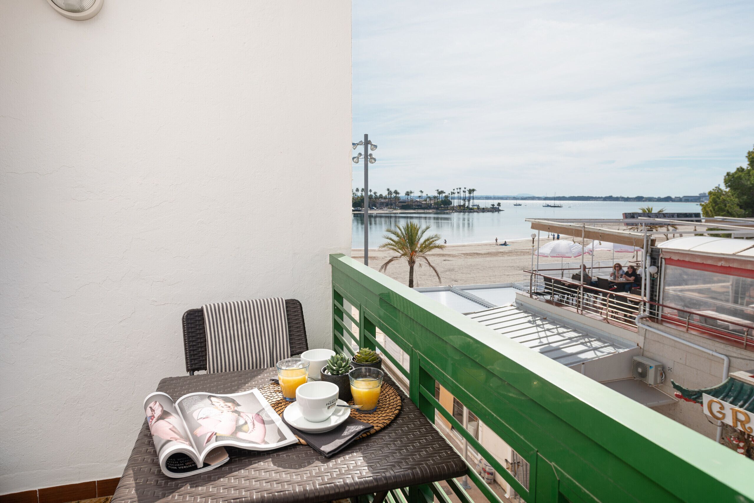 Un balcón con una mesa dispuesta para el desayuno, con vista a una playa y un puerto deportivo.